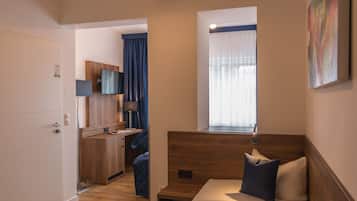 Quarto de hotel pequeno com cama, escrivaninha, cadeira, televisão e janela com cortinas.