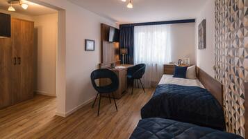 A hotel room with a wooden floor, a desk with two chairs, a bed with a dark quilt, a TV mounted on the wall, and a patterned wall.