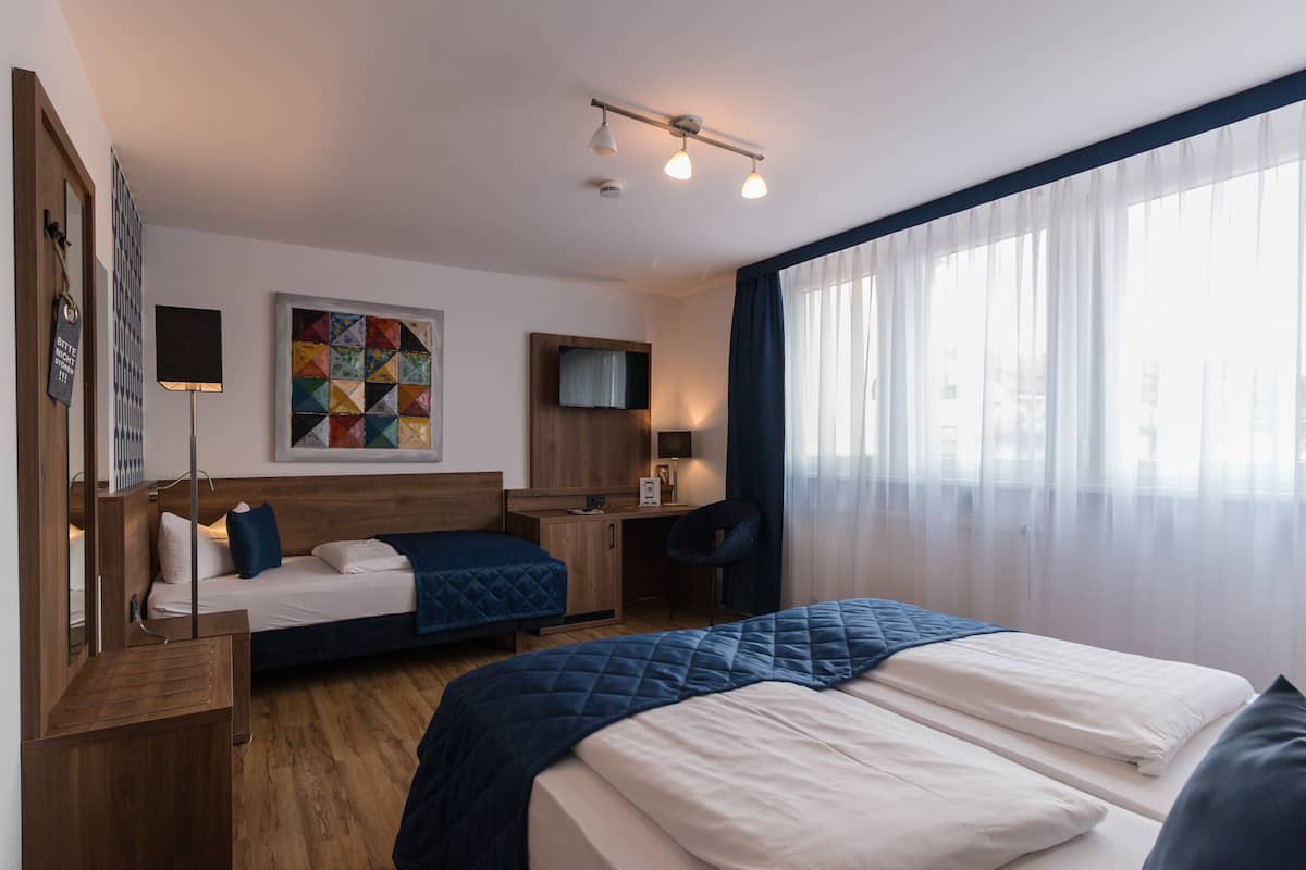 A hotel room with two beds, a desk, a chair, a TV, and a colorful quilt on the wall.