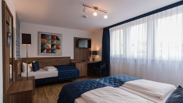 Ein Hotelzimmer mit zwei Betten, einem Schreibtisch, einem Sessel, einem Fernseher und einer farbenfrohen Stegdecke an der Wand.