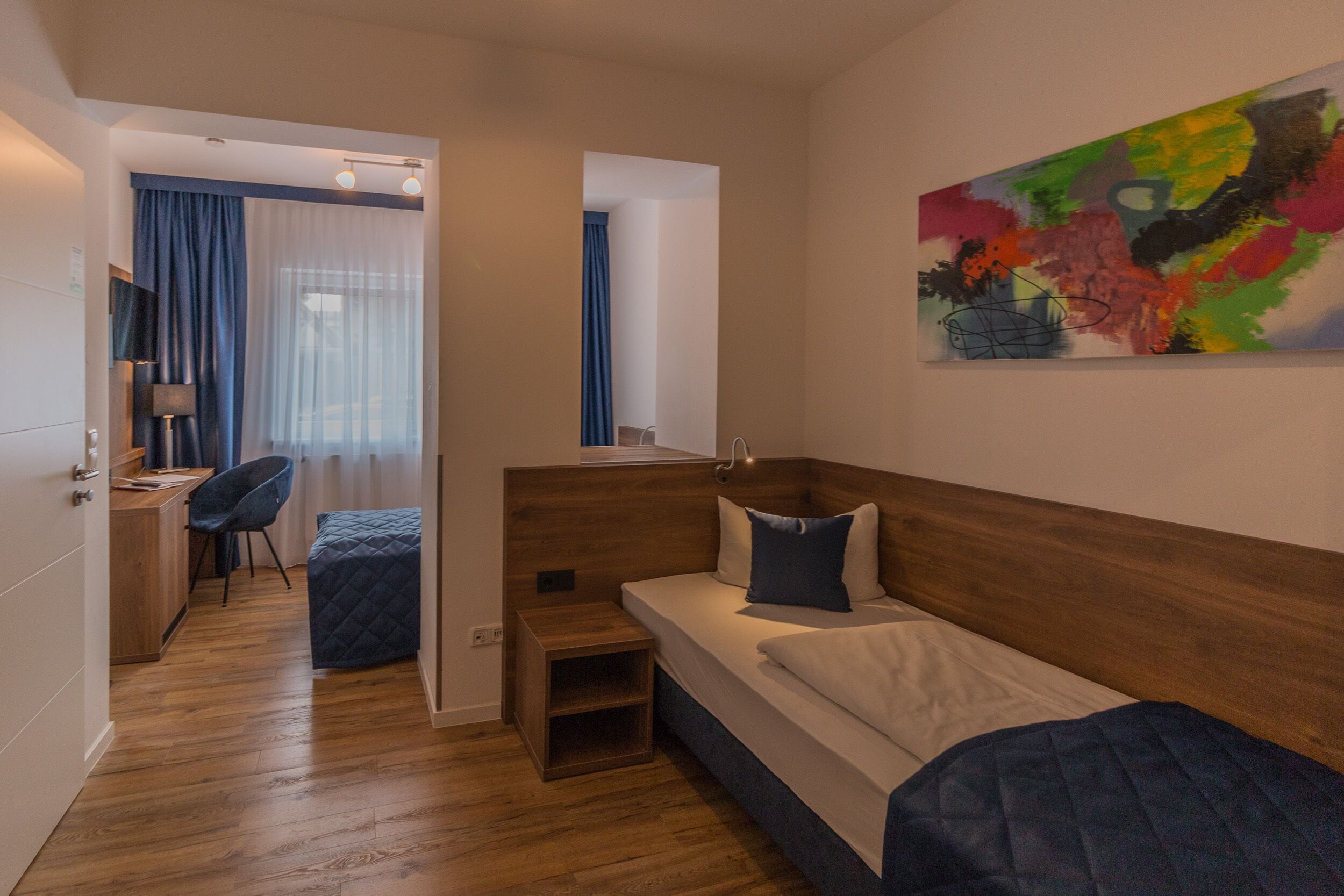 A modern hotel room with a wooden headboard, a bed with a quilted cover, a desk with a chair, a small bedside table, and a colorful abstract painting on the wall.