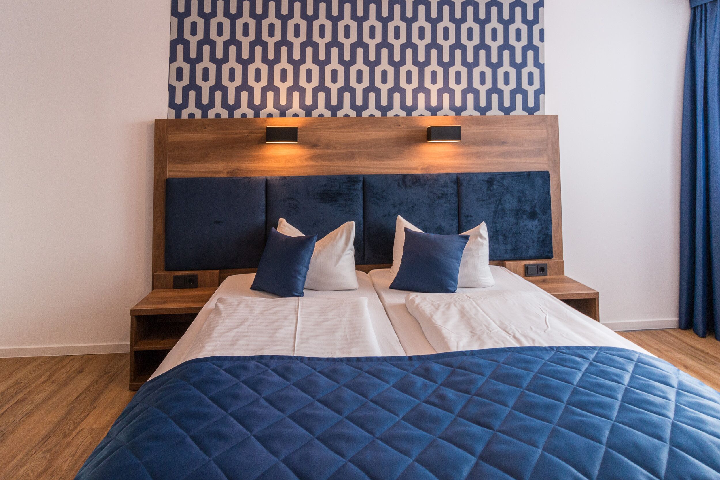 Un letto con una coperta blu, lenzuola bianche e due cuscini. Sono presenti due comodini con lampade. La parete è rivestita con carta da parati a motivi geometrici.