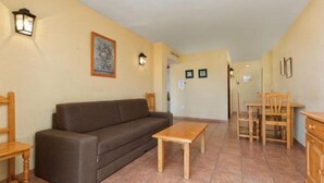 A living room with a sofa, coffee table, and wooden chairs. There are wall-mounted lights and a framed picture on the wall. - Embajador Apartamentos (Fuengirola)