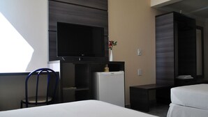 Et hotelværelse med seng, skrivebord med fladskærms-tv, lille køleskab, stol og et sengebord.