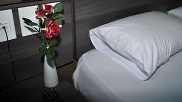 Hotellihuone, jossa on sänky, valkoiset tyynyt, valkoinen maljakko, jossa on punaisia kukkia, kaukosäädin ja seinäpistorasia.