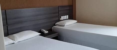 Quarto de hotel com duas camas de solteiro, cabeceira de madeira, mesa de cabeceira com telefone e ar condicionado fixado na parede.