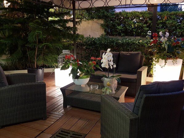 An outdoor patio area with wicker furniture, a glass coffee table, and various potted plants. - Hotel Beppe (Rimini)