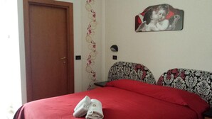 A bedroom with a red bedspread, a wooden door, patterned wallpaper, and a framed picture on the wall.