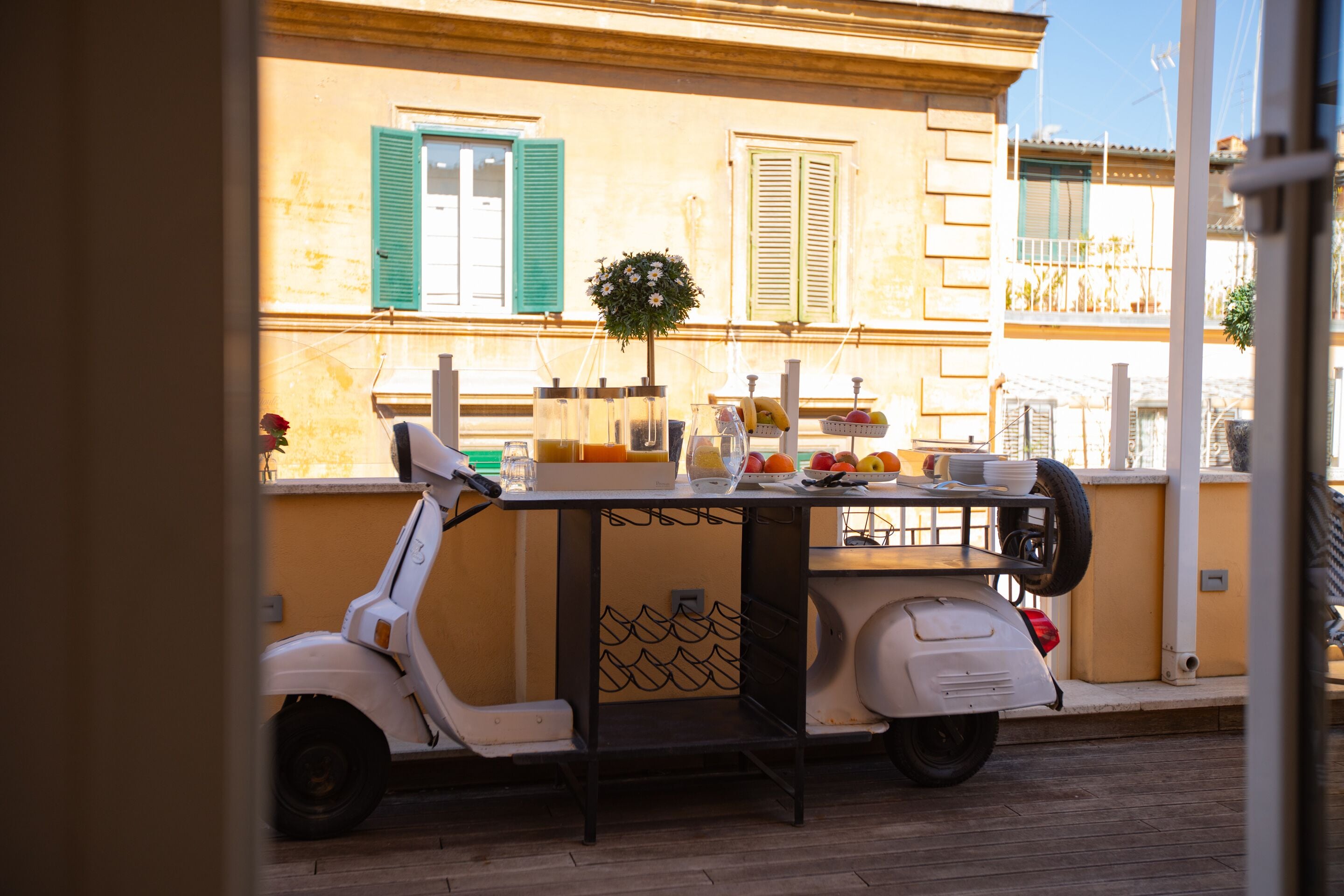 A scooter parked in front of a building with a table set up outside, featuring a basket of fruits and a potted plant.