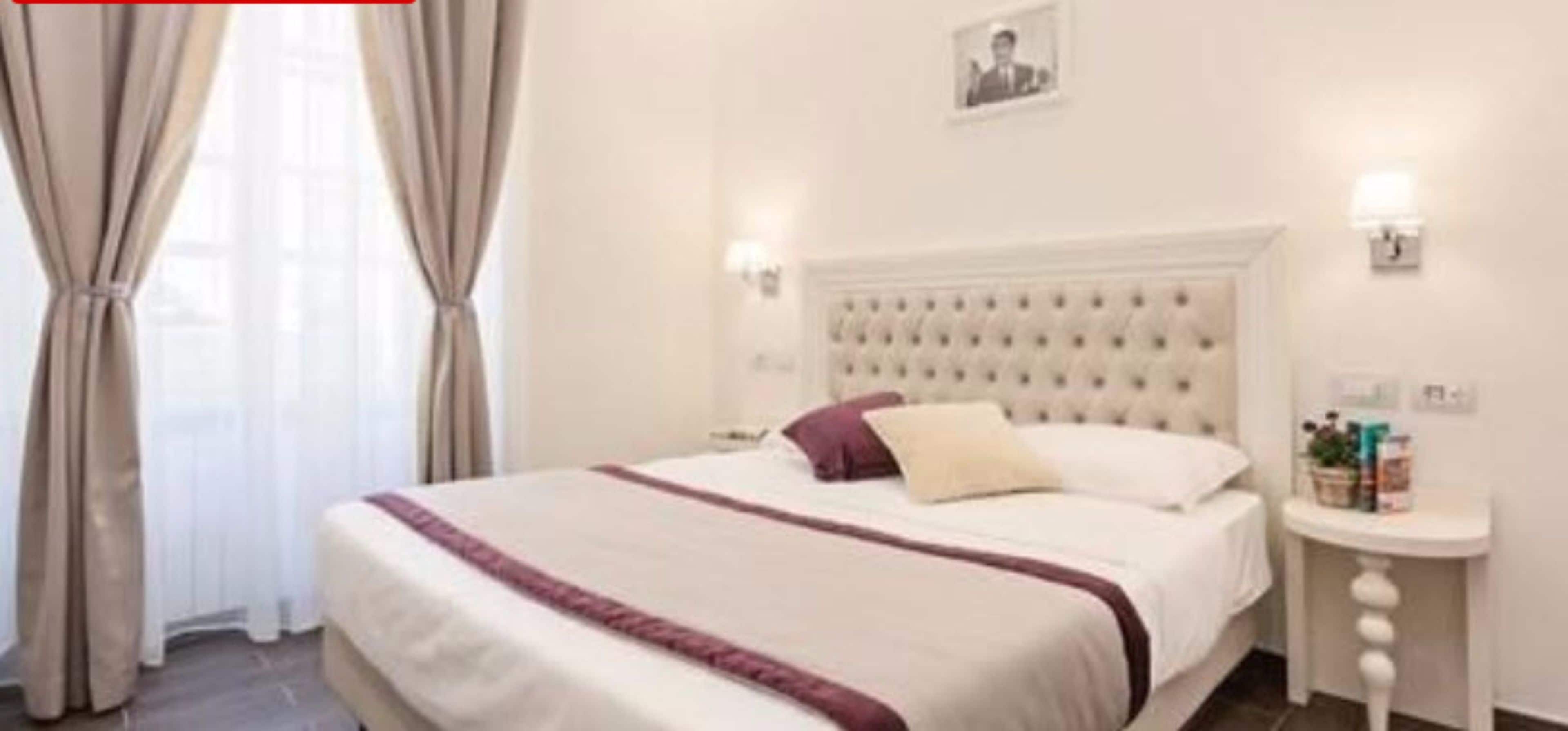 A hotel room with a large bed, a tufted headboard, two bedside tables with lamps, and a window with curtains.