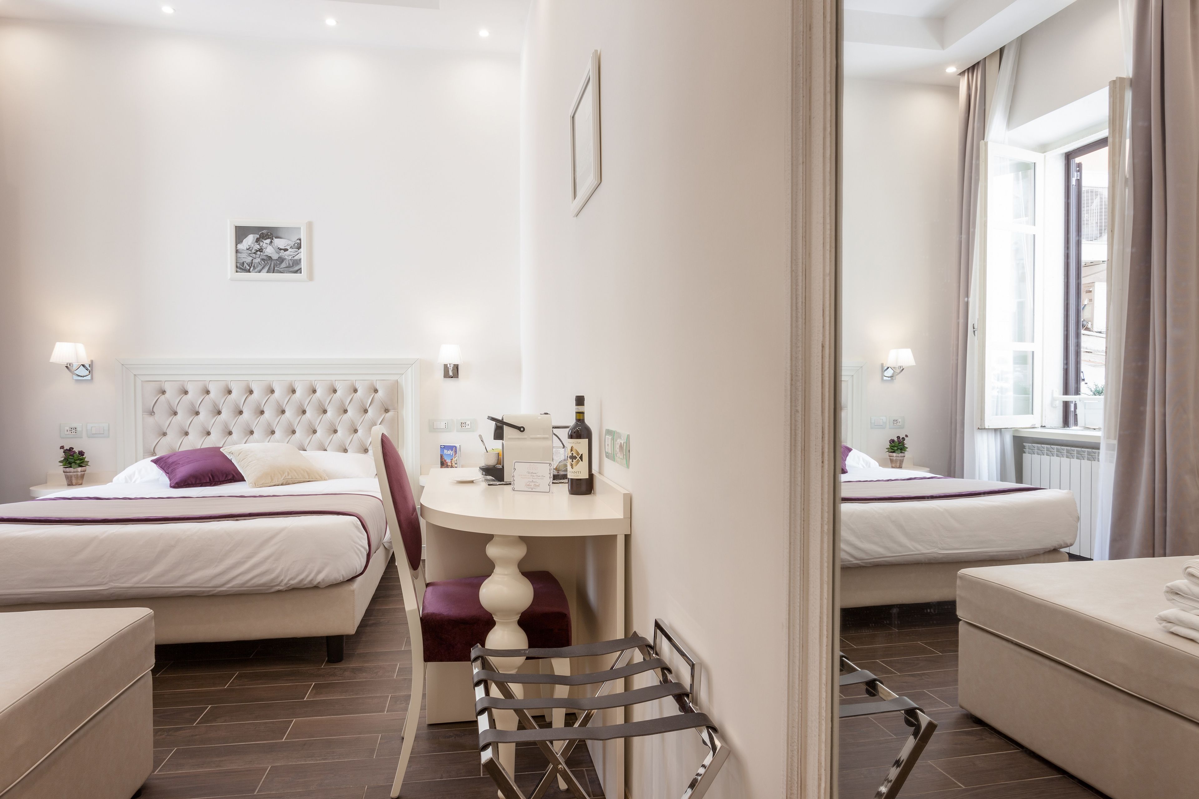 Une chambre d’hôtel avec deux lits, une petite table sur laquelle se trouvent une bouteille et des verres, une chaise, et une vue sur la rue par la fenêtre.
