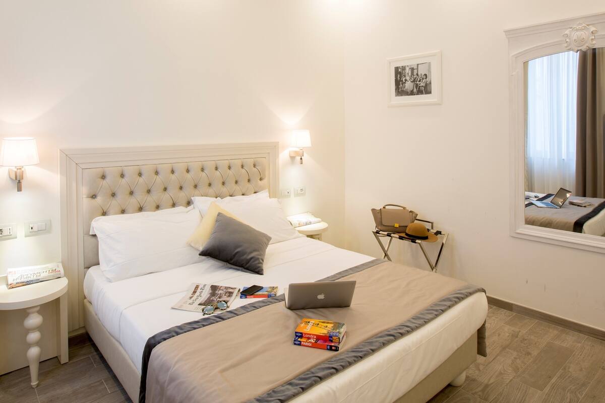 A neatly made bed with a laptop and books, a bedside lamp, a framed picture, and a window with curtains.