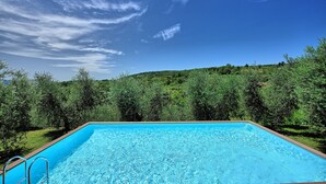 Une piscine rectangulaire à l’eau d’un bleu limpide, entourée d’une végétation luxuriante et d’arbres, sous un ciel bleu dégagé parsemé de quelques nuages.