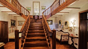 Un magnifique escalier en bois, surmonté d’un lustre, et bordé de tableaux encadrés et d’un canapé à motifs floraux.