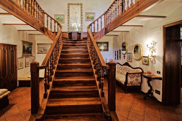 A grand wooden staircase with a chandelier at the top, flanked by framed artwork and a floral sofa.