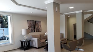 A modern living room with a sofa, a coffee table, and a chair. There are two wall-mounted paintings and a window with a view of palm trees. - Atalaia Apart Hotel (Aracaju)