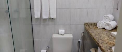 Un baño con inodoro, ducha y zona de lavamanos. Hay toallas colgadas en un perchero y una pila de toallas enrolladas en la encimera.