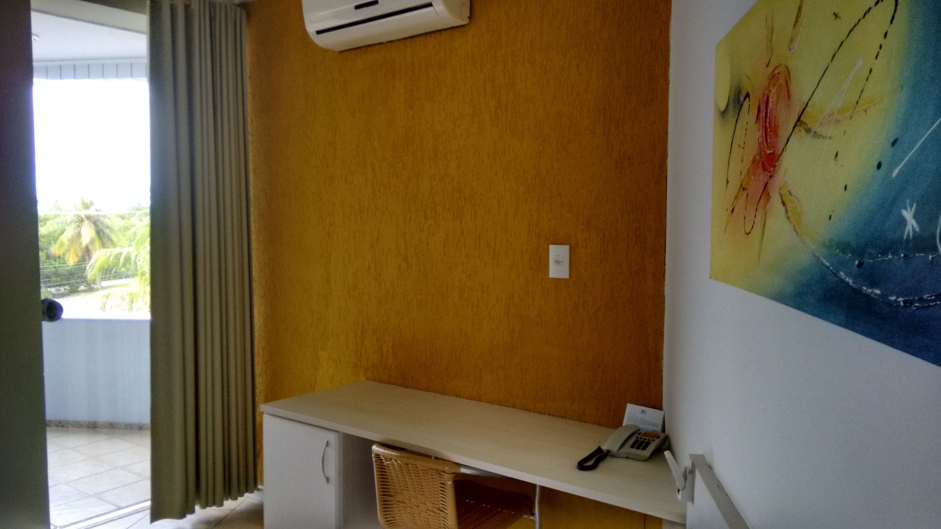A room with a yellow wall, a white desk, a wicker chair, a telephone, and a painting.