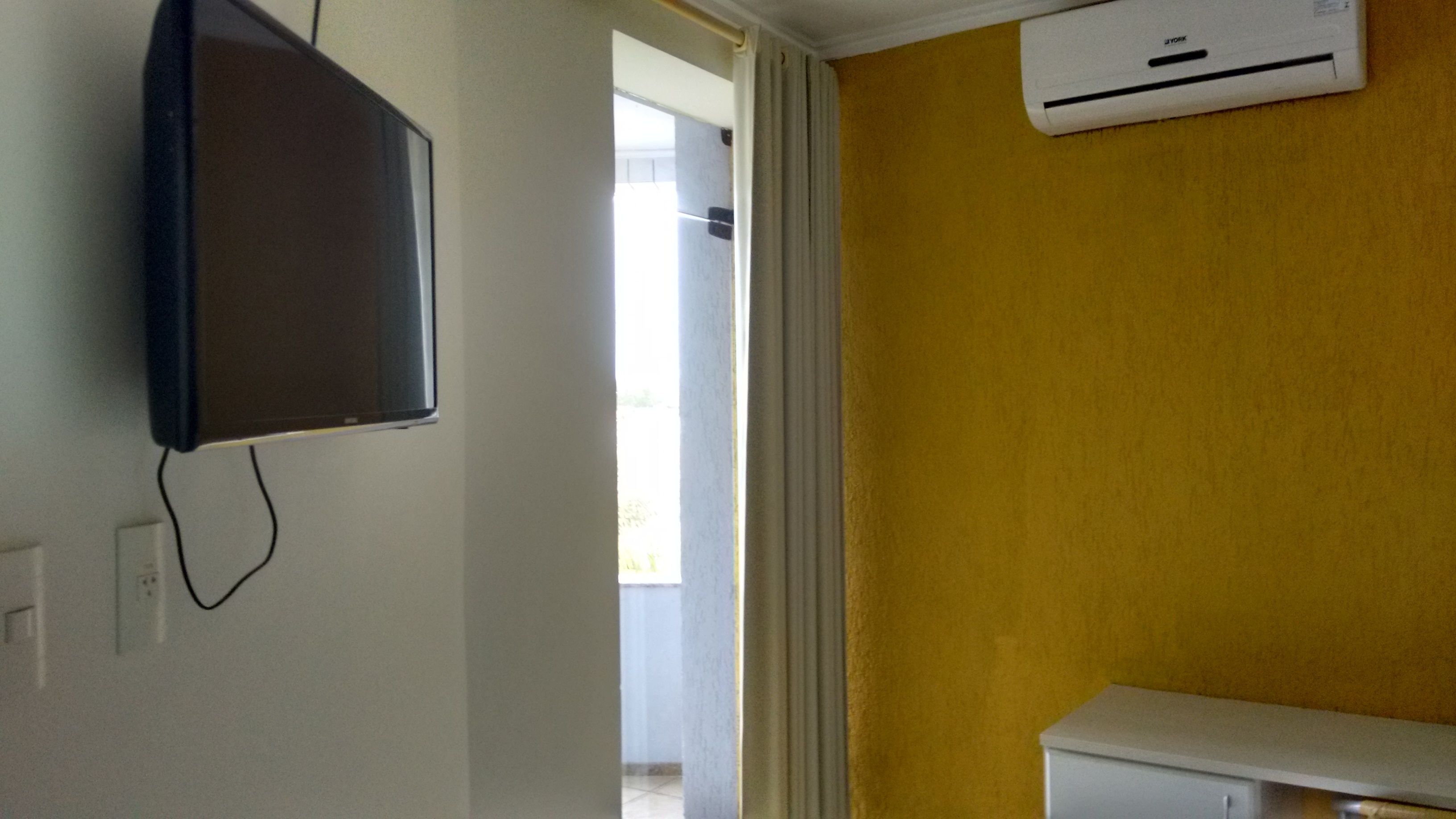 Una habitación con televisión montada, aire acondicionado, un armario blanco y una puerta que da a un área exterior.