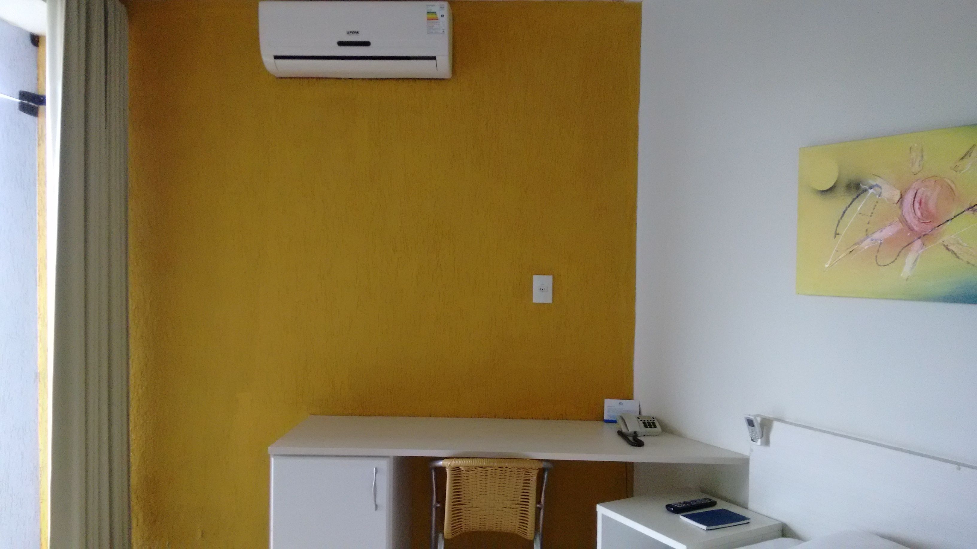 A room with a yellow wall, a white desk, a wicker chair, a white cabinet, and a painting.