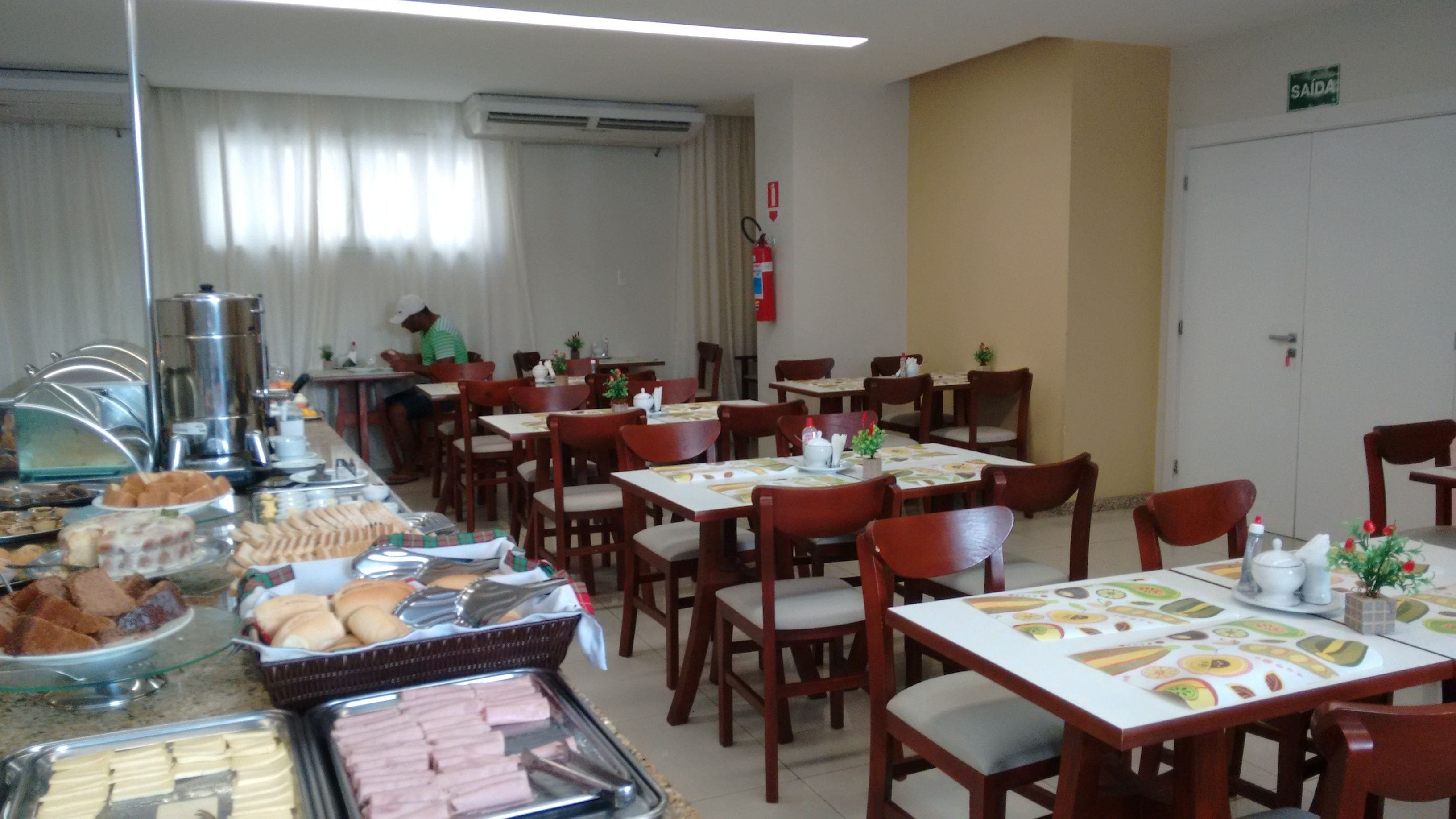 Comedor con varias mesas y sillas, bufé con distintos alimentos, y un extintor en la pared.