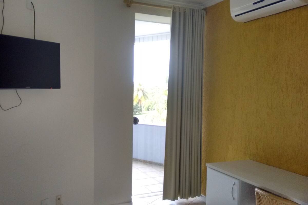 A room with a mounted TV, a window with curtains, a small table, and an air conditioning unit.