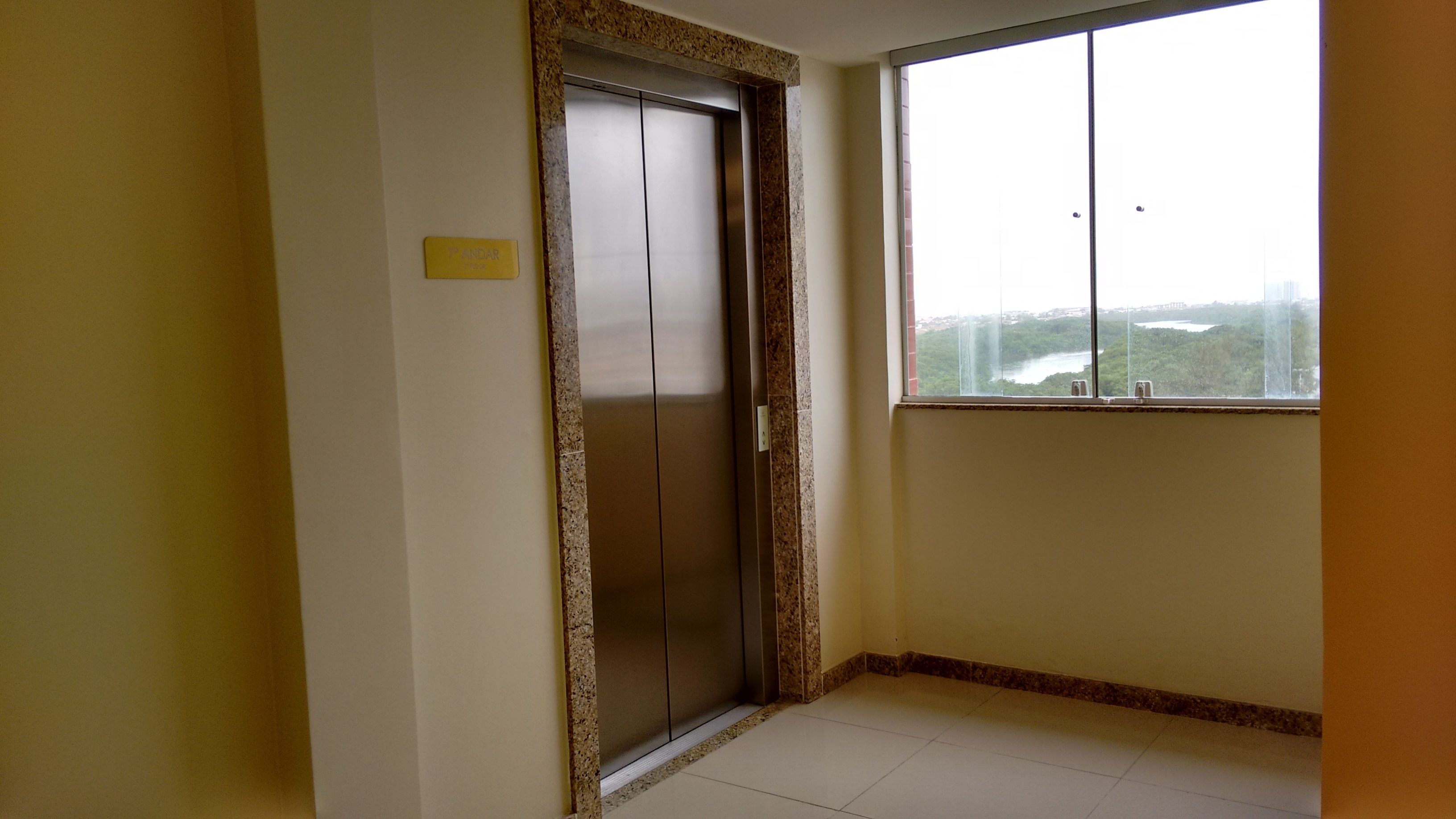 Una habitación con una puerta de ascensor cerrada, una ventana que ofrece vistas a un río y follaje, y un cartel amarillo en la pared.