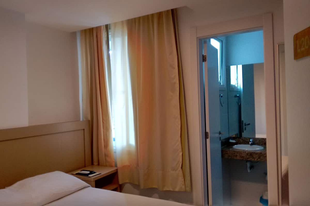 A hotel room with a bed, a small bedside table, a window with curtains, and a bathroom visible through an open door.