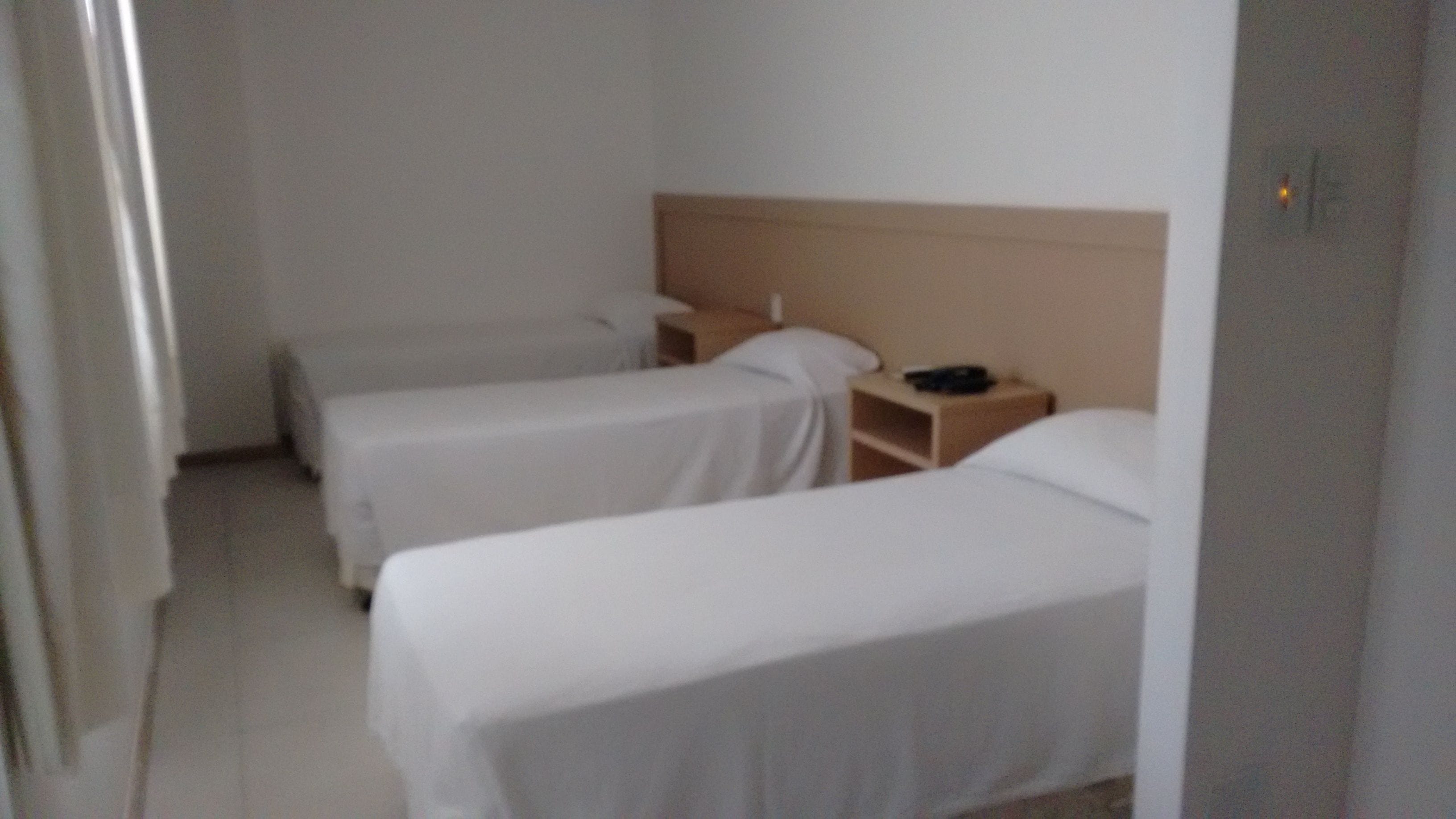 Quarto de hotel com duas camas de solteiro, cabeceira de madeira, mesa de cabeceira com abajur e uma pequena prateleira.
