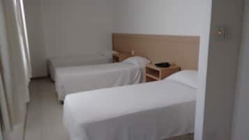 Quarto de hotel com duas camas de solteiro, cabeceira de madeira, mesa de cabeceira com abajur e uma pequena prateleira.
