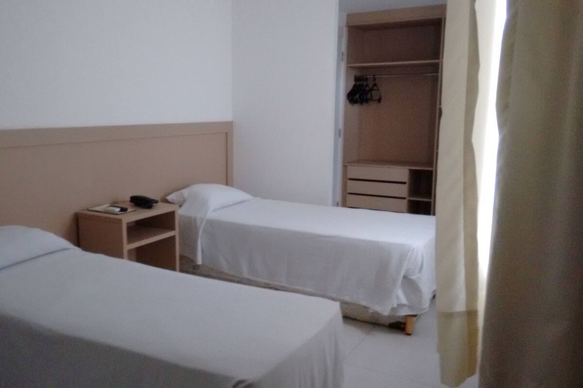 A small, clean room with two single beds, a bedside table with a phone, and a closet with hanging clothes.