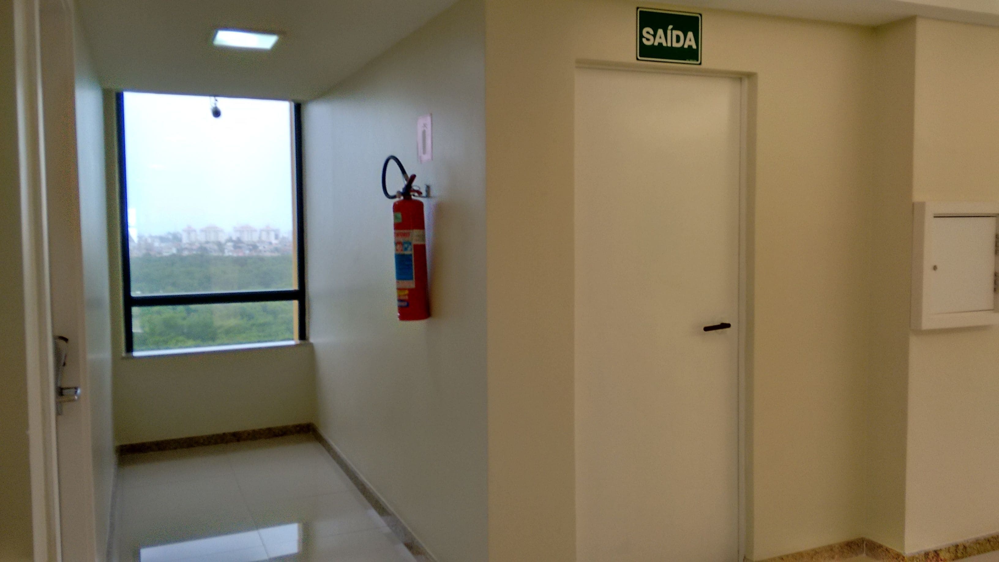 Un pasillo con una puerta que indica "SAÍDA" (salida), un extintor y una ventana con vistas a edificios.