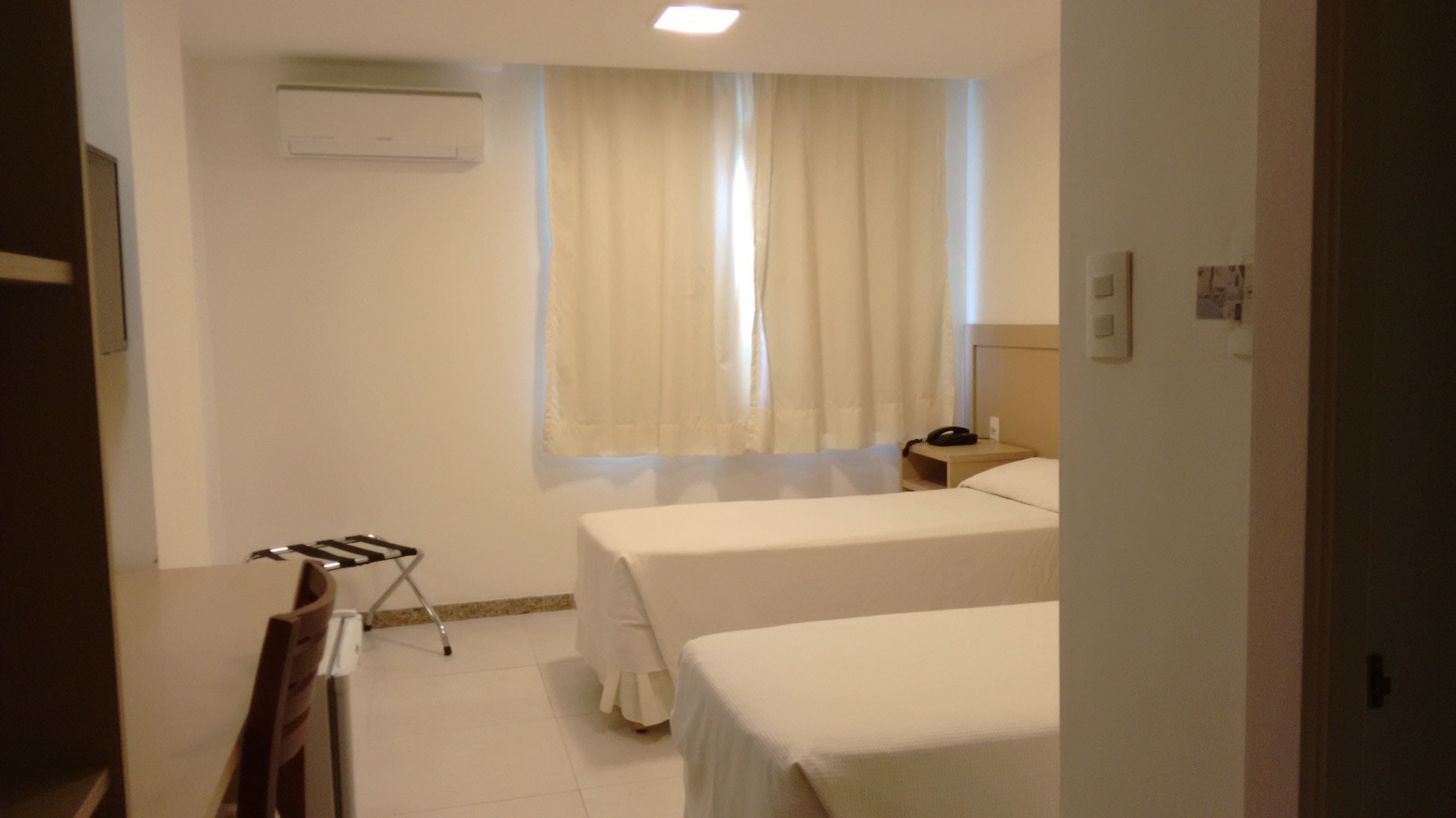 A hotel room with two beds, a bedside table with a lamp, a window with curtains, and an air conditioning unit on the wall.