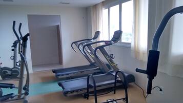 Een sportschool met loopbanden, een hometrainer en een roeimachine. Er is een raam met gordijnen en een deur naar een andere ruimte.