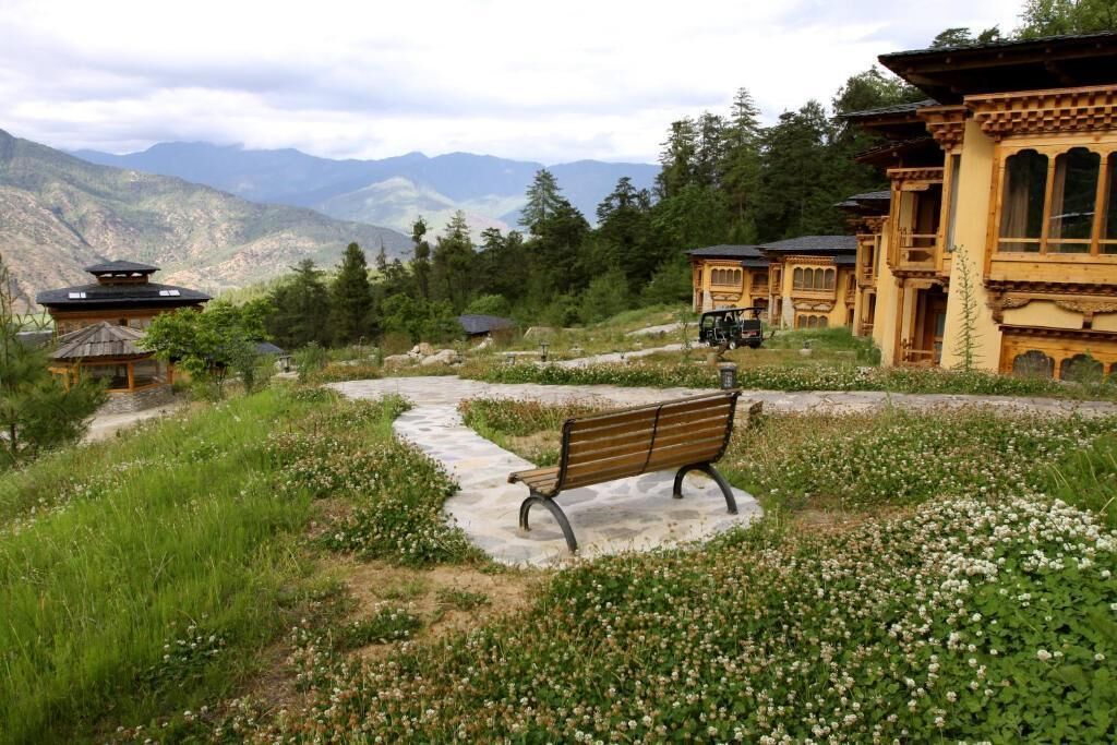 A traditional wooden building with a stone pathway leading to a bench surrounded by greenery and flowers.