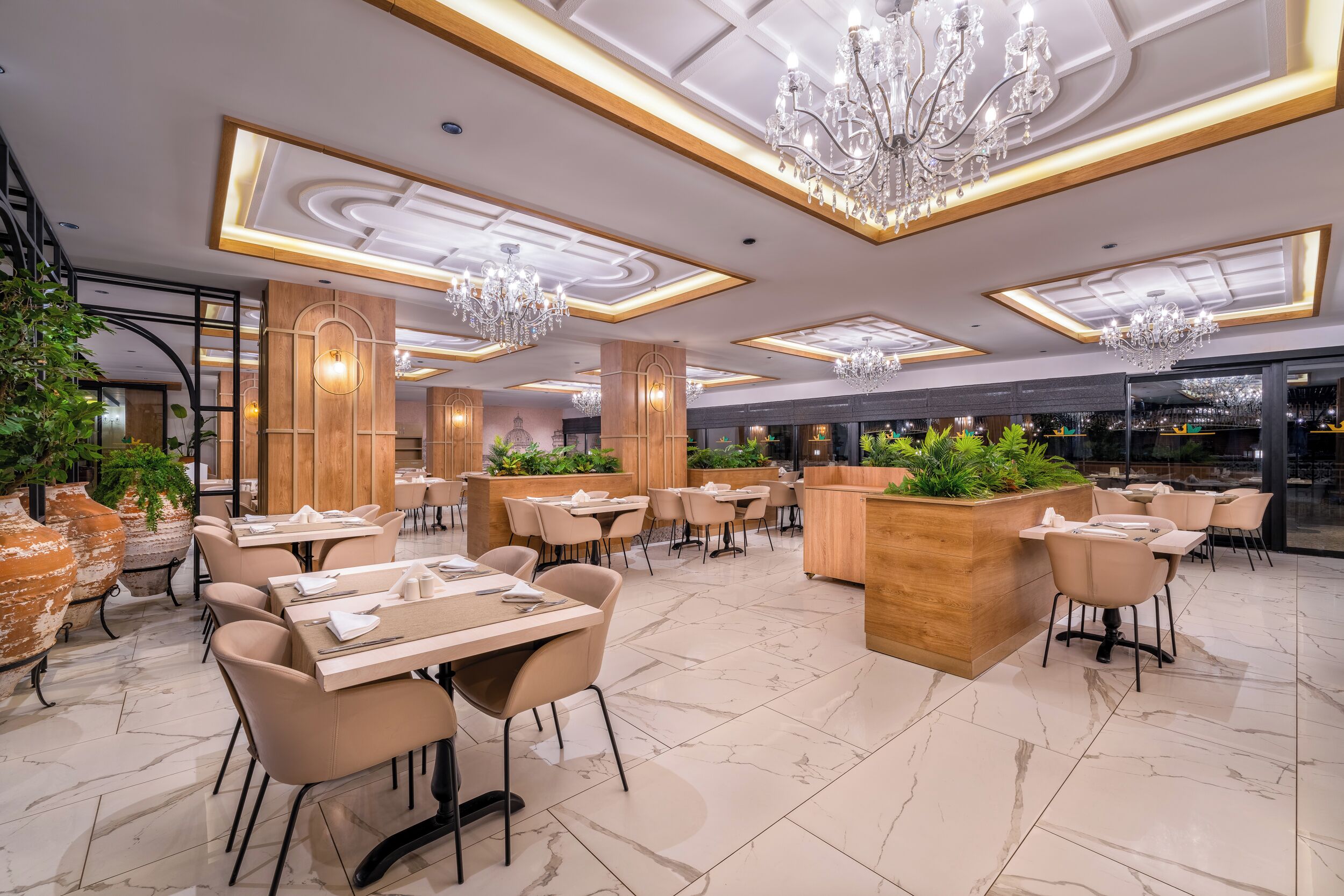 Un restaurant spacieux doté d’un sol en marbre, de touches de bois et de plusieurs lustres. Les tables sont disposées avec soin et accompagnées de chaises, et des plantes en pot apportent une touche de verdure.