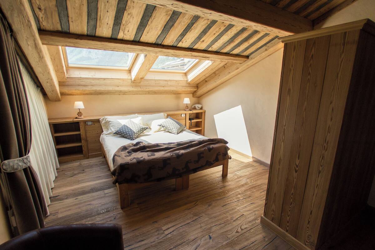 Una camera da letto con soffitto in legno, lucernari, un letto con cuscini, un comodino e un tavolino di fianco al letto.