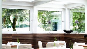 A modern restaurant with large windows, white furniture, and a wooden accent wall. - Hotel Bonjour (Bad Soden am Taunus)