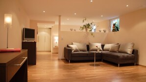 A modern living room with a grey sectional sofa, wooden flooring, a TV on a stand, and a plant on a table. - Hotel Bonjour (Bad Soden am Taunus)