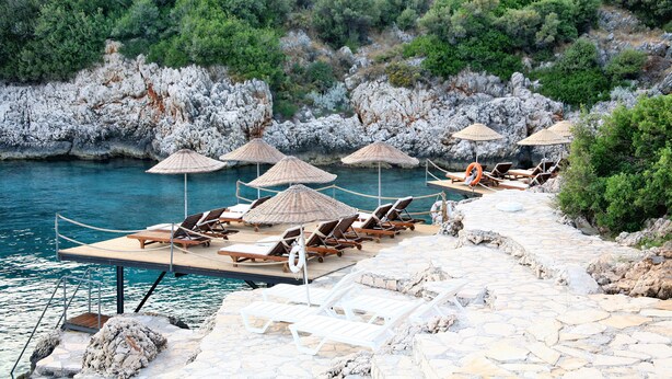 A stone deck with lounge chairs and umbrellas by a body of water, surrounded by rocky terrain and greenery. - Kulube Hotel (Kas)