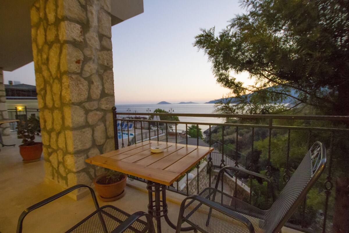 A balcony with a wooden table and metal chairs, overlooking a body of water and greenery.