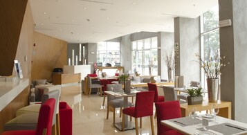 A modern restaurant with a mix of red and grey chairs, a wooden reception desk, and large windows providing natural light.