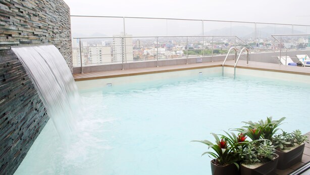 Una piscina en la azotea con cascada, rodeada por una barandilla de cristal y plantas en macetas.