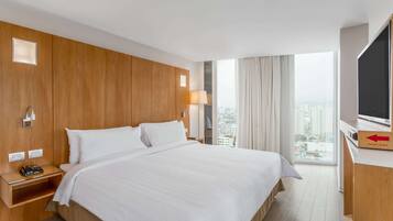 A hotel room with a large bed, wooden paneling, a TV, a desk with a computer, and a window with a city view.