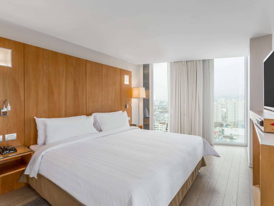 A hotel room with a large bed, wooden paneling, a TV, a desk with a computer, and a window with a city view.