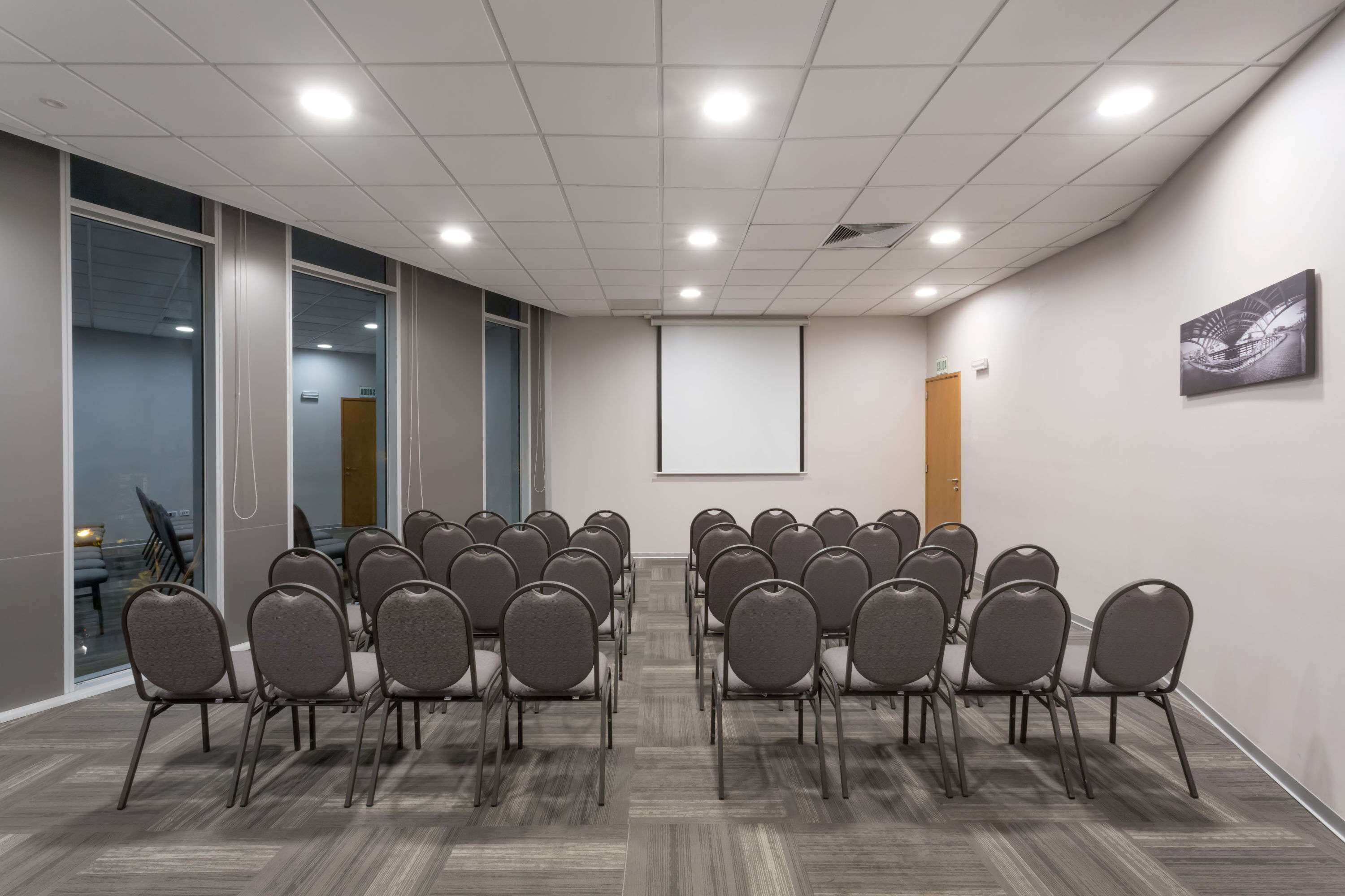 Uma sala de conferências com fileiras de cadeiras, uma tela de projetor e um grande janelão.