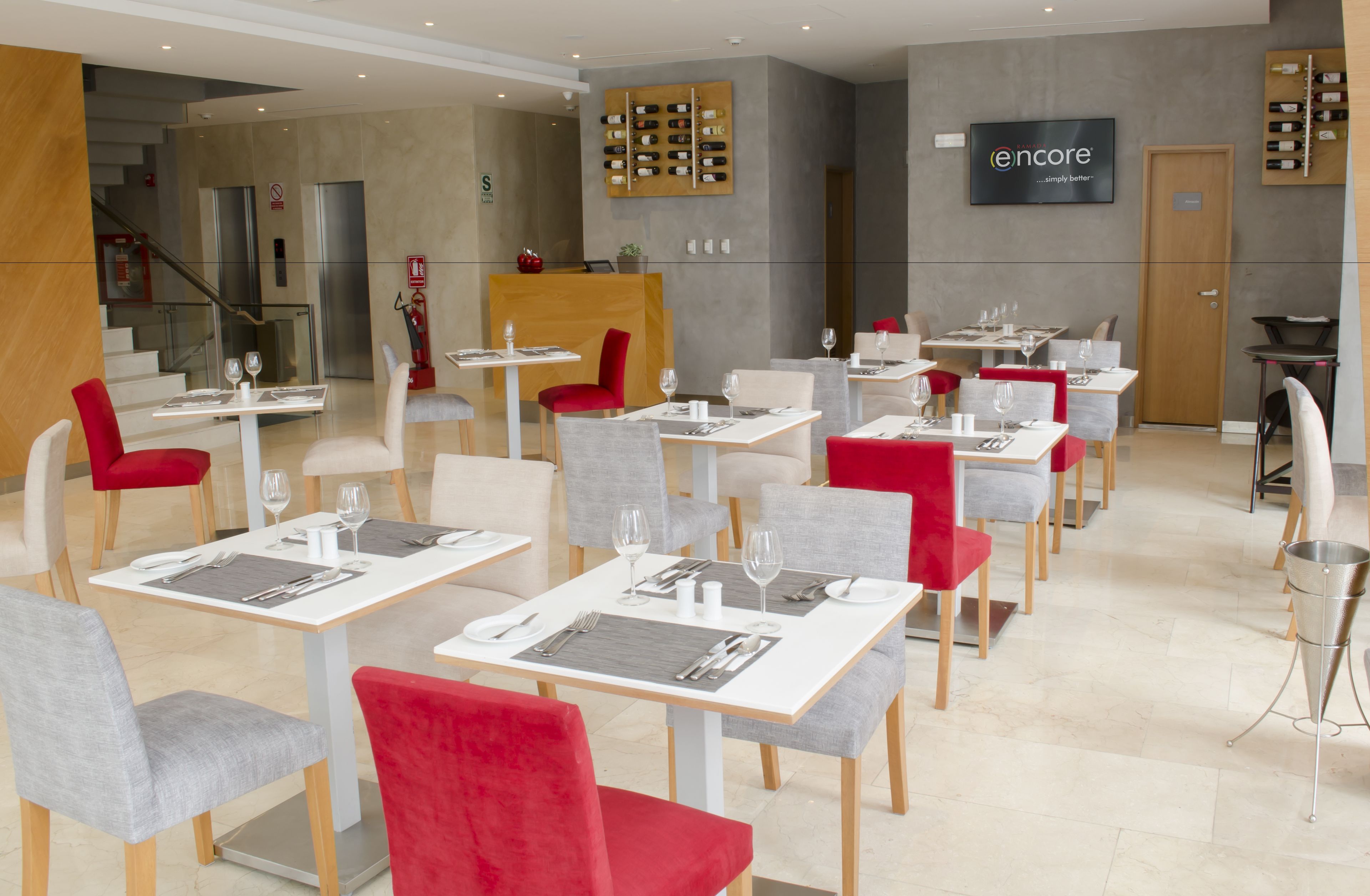 Um restaurante moderno com mesas brancas e vermelhas, cadeiras cinzentas e um display de vinhos na parede.