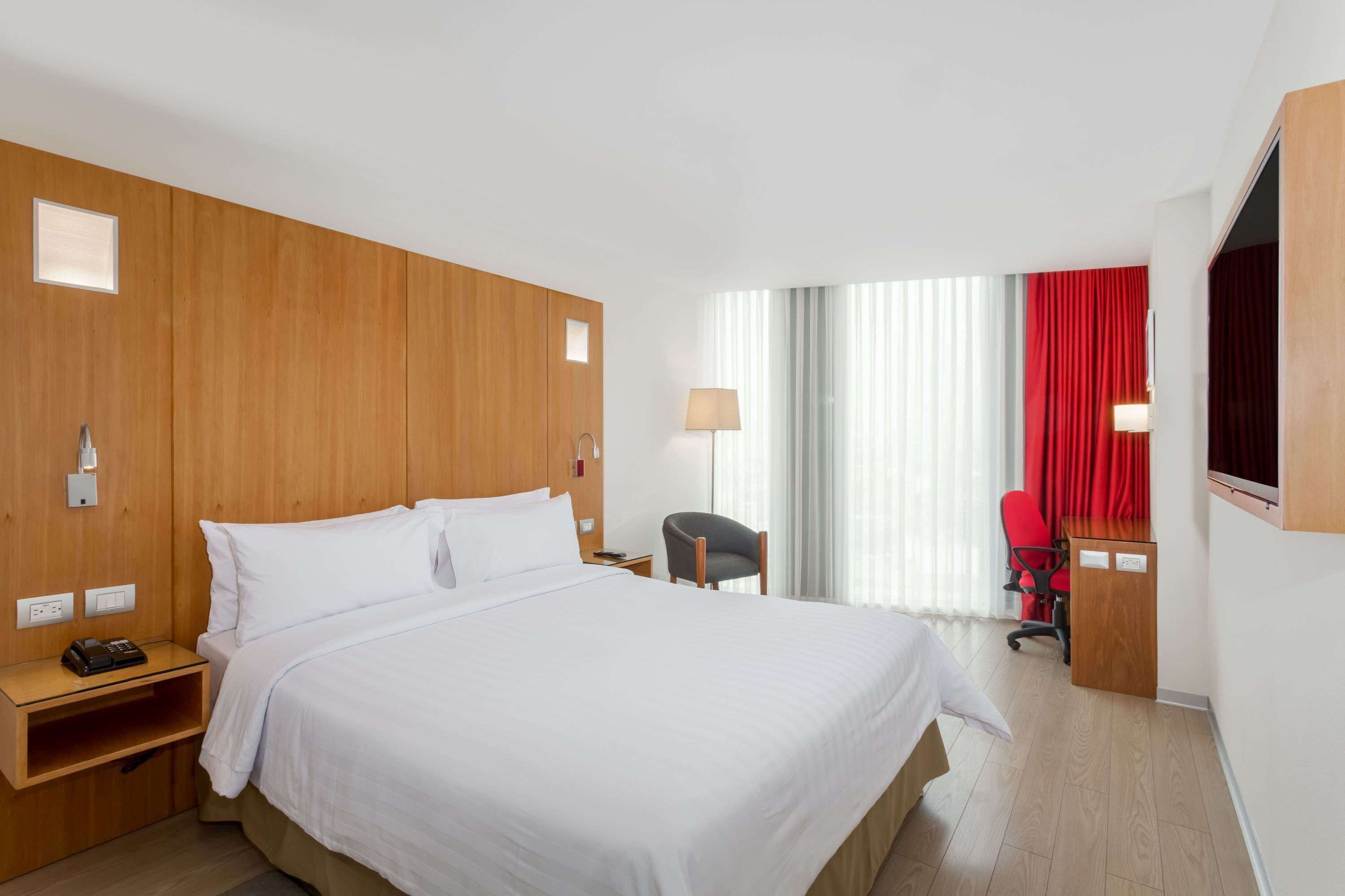 A hotel room with a large bed, wooden headboard, a desk with a chair, a lamp, and a window with curtains.