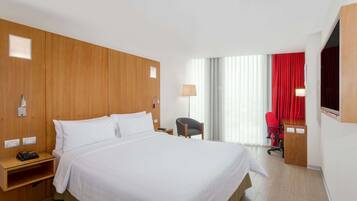 A hotel room with a large bed, wooden headboard, a desk with a chair, a lamp, and a window with curtains.