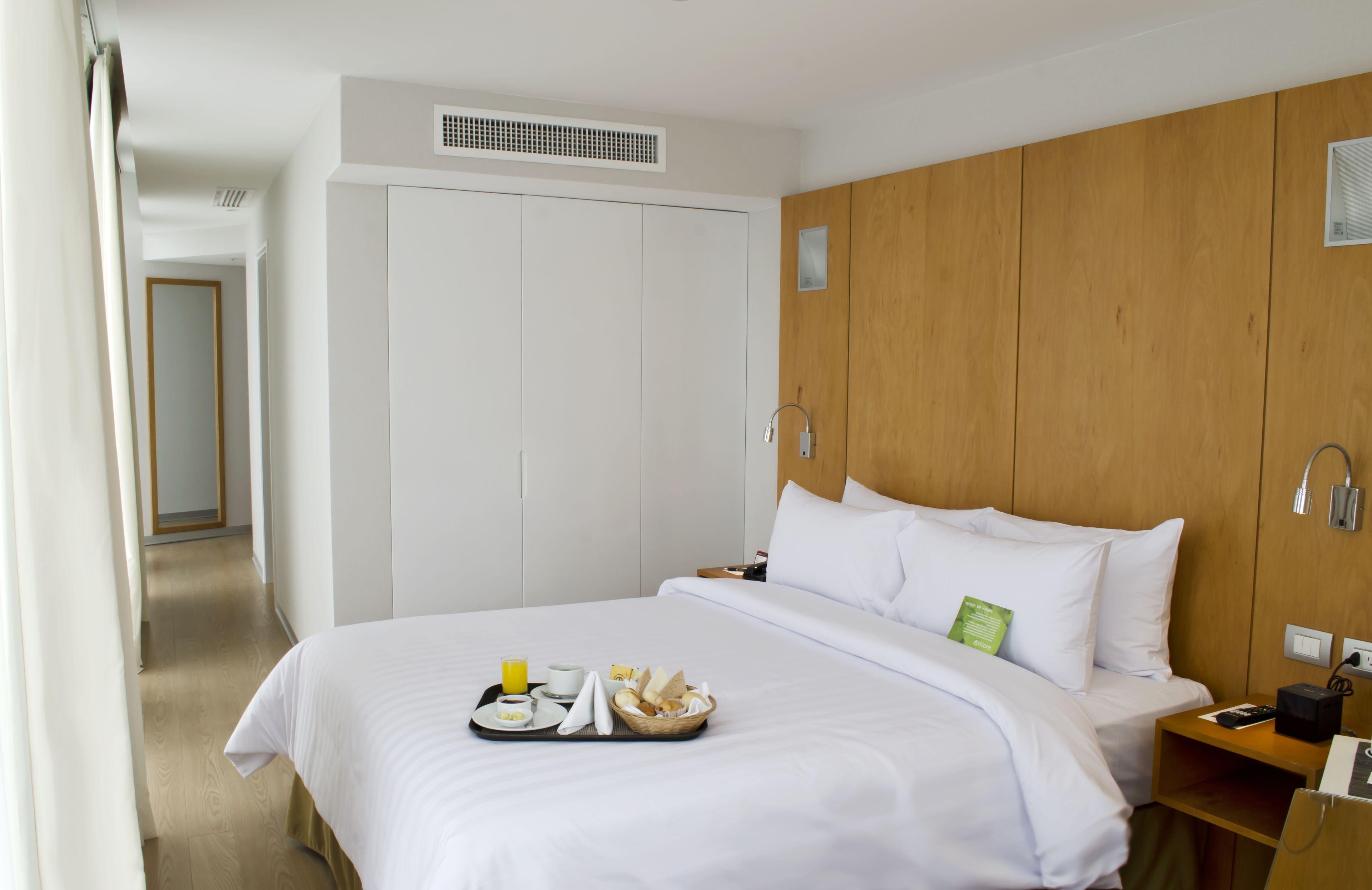 A hotel room with a bed, a wooden wardrobe, a bedside table with a phone, and a tray with breakfast items.